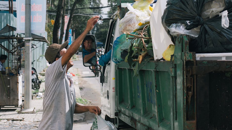 A side view of a medium-sized waste collection truck parked on a street in front of a residential building with multiple windows and balconies, surrounded by leafless trees. The truck is designed for