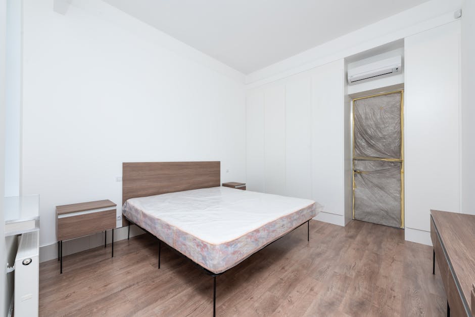 A minimalistic bedroom with white walls and ceiling, featuring a double bed with a light wooden headboard and matching bedside tables on each side, both with thin black metal legs. The bed has a plain