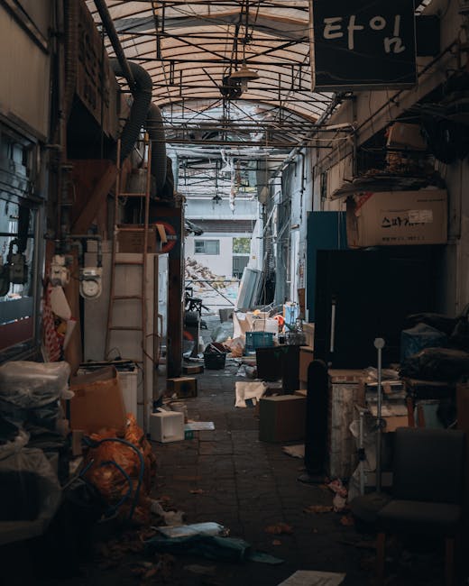 The interior of a large, dimly lit room with a high ceiling shows a significant accumulation of mixed household rubbish scattered across the floor. The debris includes plastic bags, crumpled paper, br