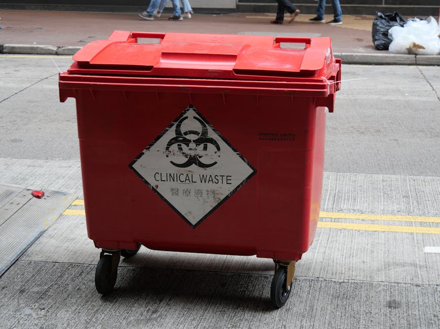 A worker wearing a yellow waterproof suit with a hood, black gloves, and safety goggles is engaged in hazardous waste handling outdoors. The worker is leaning over a large black plastic bucket, partia